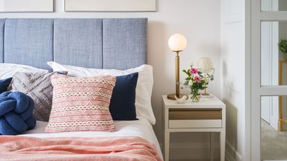 Learn how to create a hotel-style bedroom. Here is a bedroom with a light blue bed with white sheets and pink and blue throw pillows and blankets, and a white nightstand next to this with a tall gold lamp and a vase with flowers in it