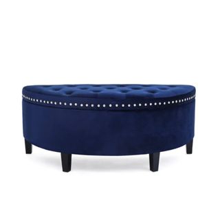 A half oval shaped navy blue storage ottoman with a tufted button top, metal studs on the edge, and black legs