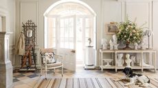 Bright and airy entryway with a striped rug on tiled patterned floor, a black and white spaniel, open glass-panelled white door, wooden umbrella stand and wooden sideboard covered in busts and flowers. The walls are white and panelled.
