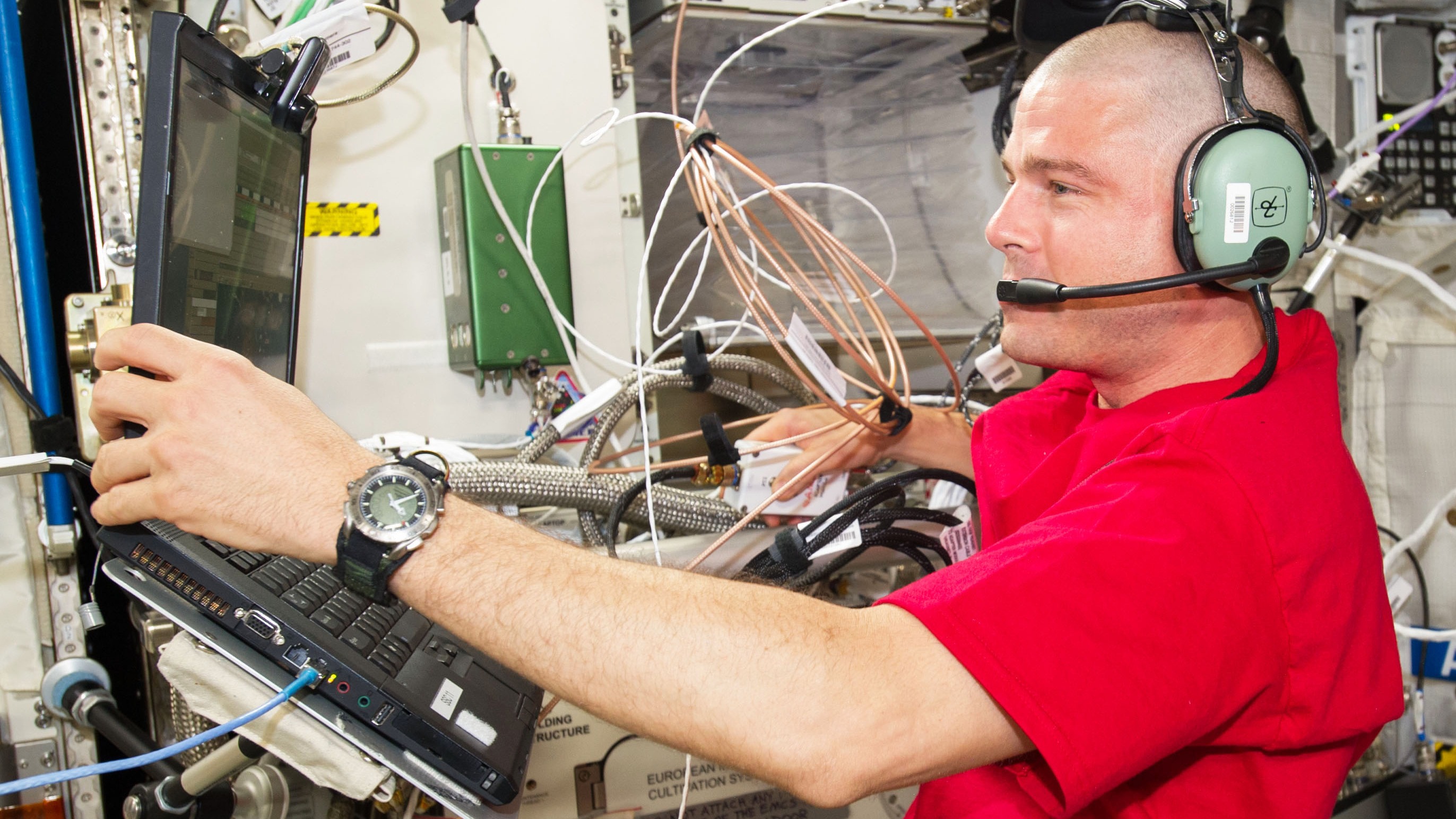 Reed Wiseman dans un casque sur la Station spatiale internationale.  Il tient un ordinateur portable sur un support avec sa main gauche