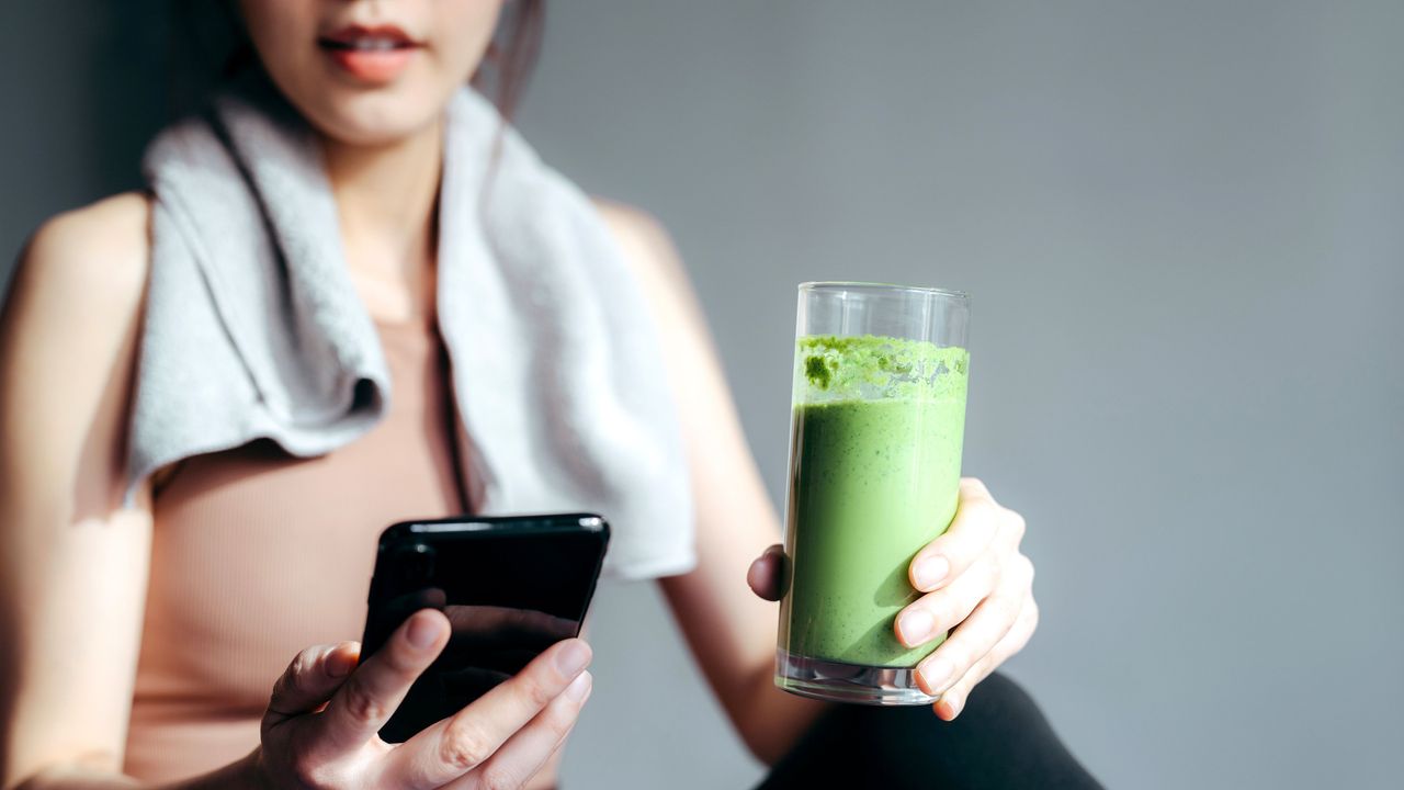 close up of confidence young asian sports woman taking a break, refreshing with healthy green juice and using smartphone after fitness work out exercising practicing yoga at home in the fresh bright morning