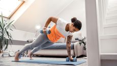 A woman performs a renegade row with light dumbbells on a yoga mat in a living room. She is in a push-up position, with one dumbbell on the mat and the other bent at the elbow holding the dumbbell near her hip. Behind her we see a couch, alarm clock and plant.