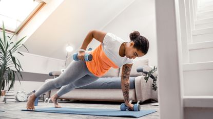 A woman performs a renegade row with light dumbbells on a yoga mat in a living room. She is in a push-up position, with one dumbbell on the mat and the other bent at the elbow holding the dumbbell near her hip. Behind her we see a couch, alarm clock and plant.