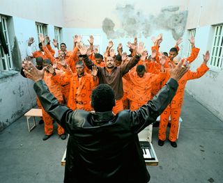 Sunday church service at Beaufort West Prison - There is a man wearing a black jacket standing in front of prisoners dressed in orange. Everyone has both arms up in the air