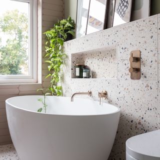 bathroom with large contemporary bath, terrazzo tiles and rose gold brassware, with a shelf featuring framed pictures and a trailing houseplant