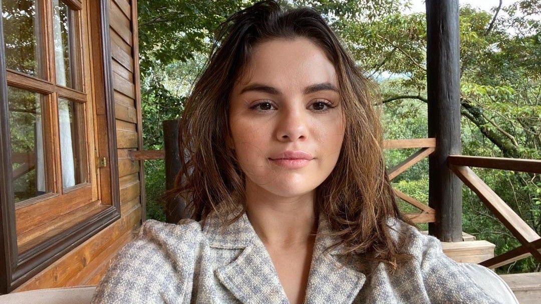 Selena Gomez sits in front of a cabin without makeup while wearing patterned pajamas