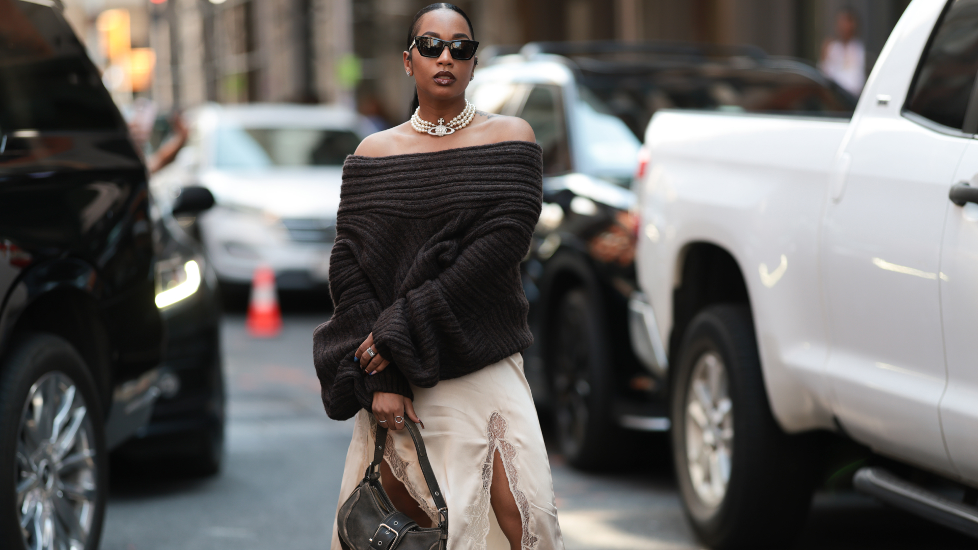 Fashion week guest is seen wearing an off-the-shoulder dark brown chunky knit sweater over a light cream-colored silky slip dress with a high slit and lace trim. She carries a brown leather handbag with a buckle in her left hand, while her right hand features rings and blue-toned nail polish. Accessories include a pearl Vivienne Westwood necklace and large black sunglasses, with her hair slicked back on Lapointe Show September 13, 2025 in New York City.