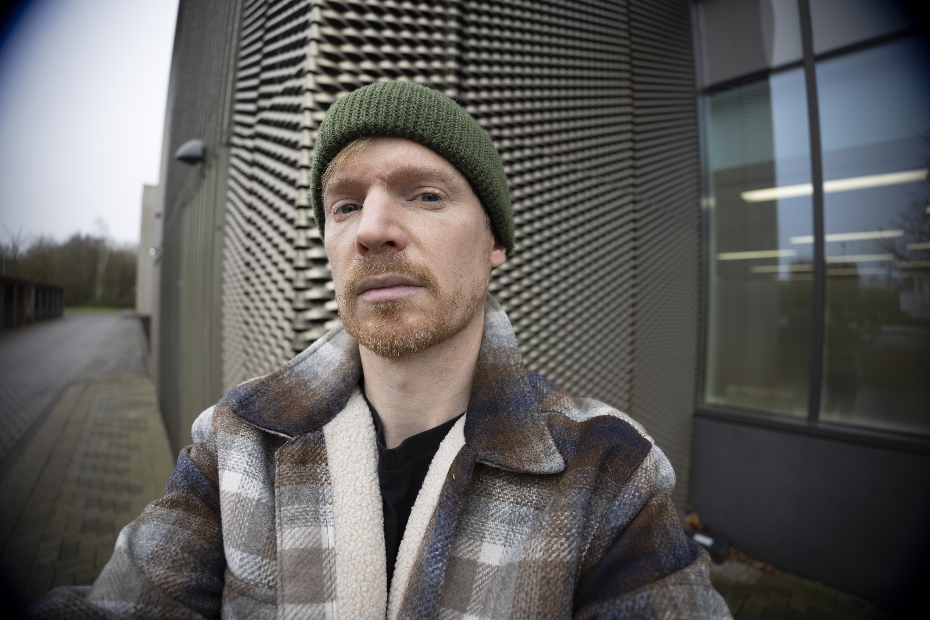 A man in a green beanie and shacket's selfie, outdoors in gray urban area