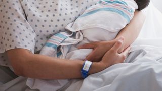 close up photo of a mother's arms holding a baby; the woman's skin is tan and she's wearing a hospital bracelet on her wrist and a hospital gown