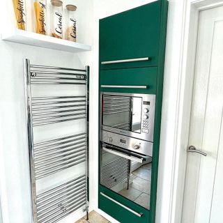 corner of a kitchen containing double inbuilt stoves stacked on top of each other with green cupboards above and below, next to a wall rail radiator and a shelf containing jars of spaghetti and cereal