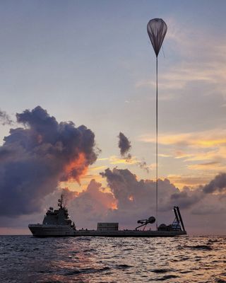 A long boat with flat mid-deck floats on the ocean against a cloudy sky at dawn or dusk. A long, towering cable stretches from a sphere resting on the back deck, upward to the sky connected to a large floating balloon.