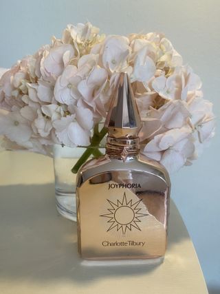 a bottle of charlotte tilbury joyphoria perfume in front of a vase of hydrangea flowers and on a table