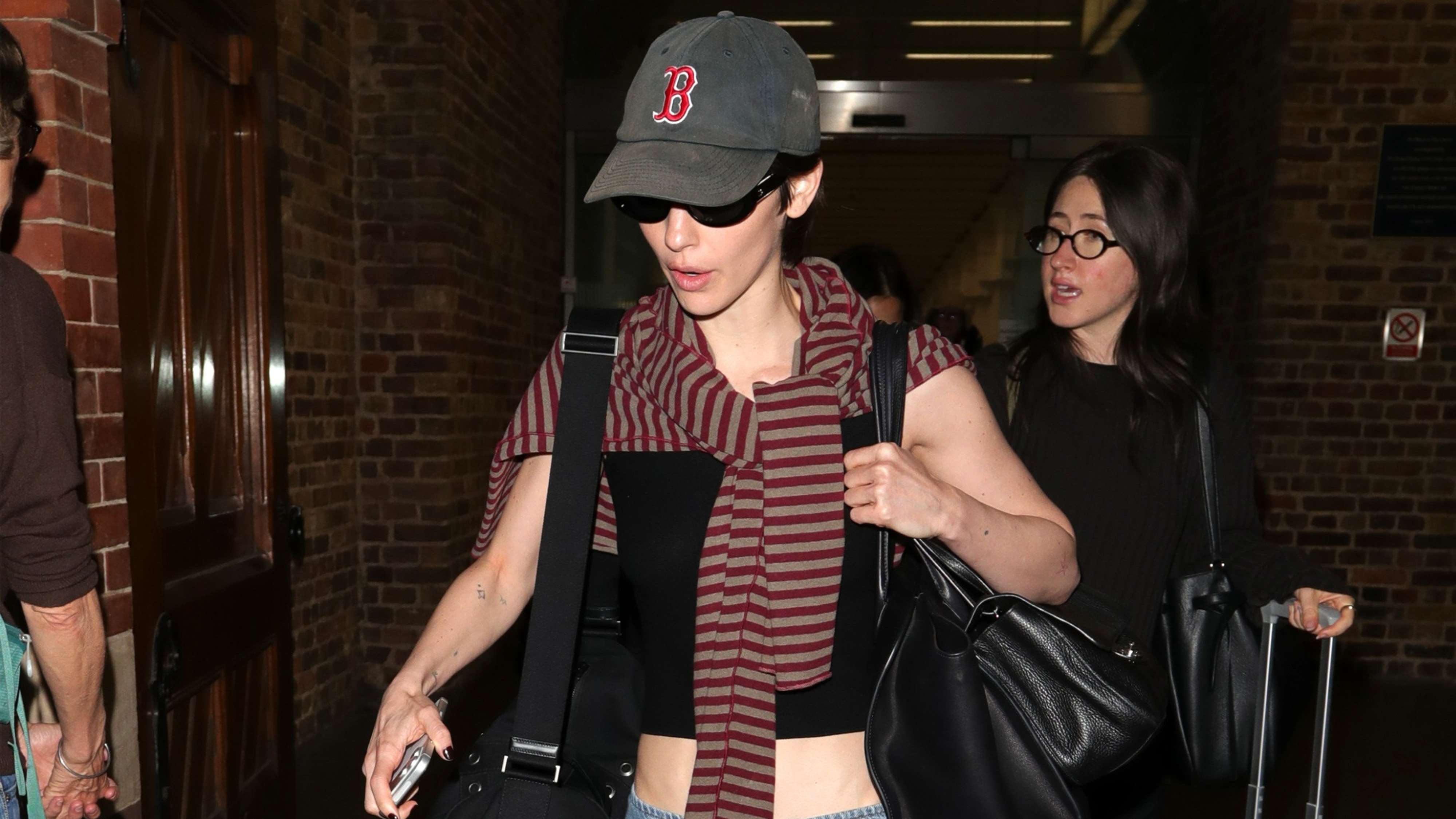 Gracie Abrams wears a Boston hat, red stripe sweater, and cropped black tank top.