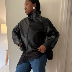 Woman wearing funnel neck leather jacket, lace mini dress, and blue jeans.