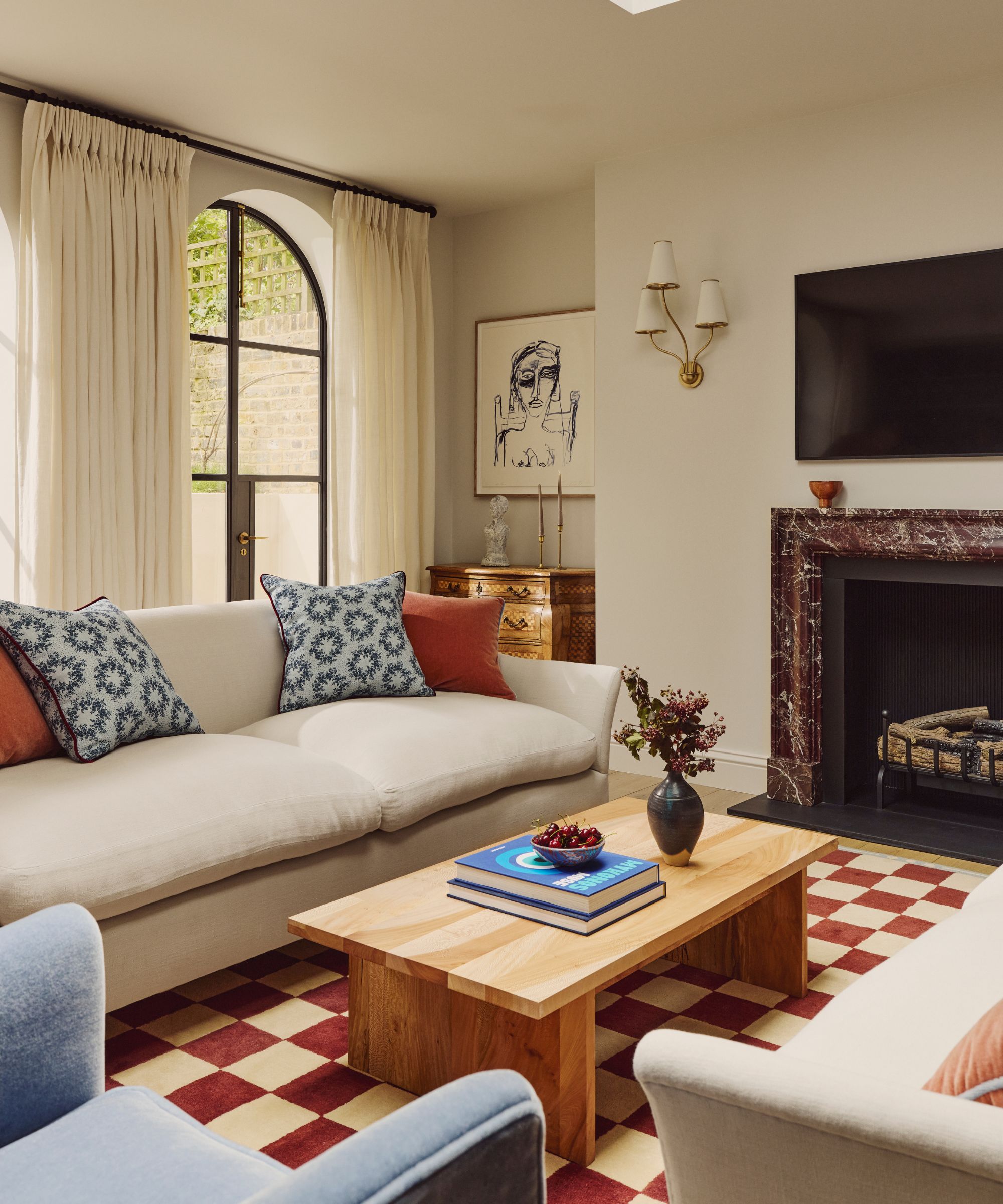 neutral living room with a red marble fireplace surround, neutral sofa with blue and red cushions, blue accent chair, and red and white checkerboard rug with a wooden coffee table
