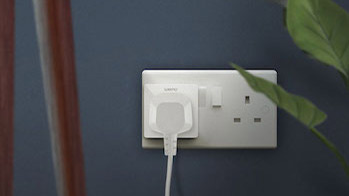 WeMo WiFi smart plug connected to a power outlet on a blue wall with a table and plant next to it