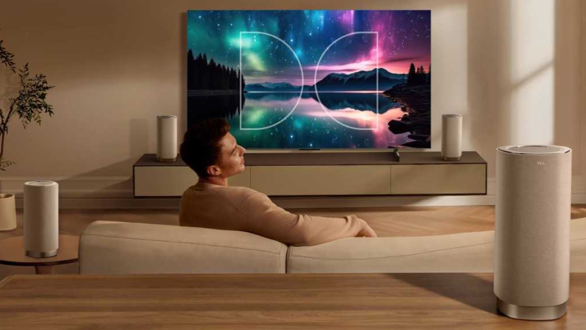 TCL Z100 surround speakers in a beige TV room behind a beige sofa on which sits a man. Guess what colour he's wearing.