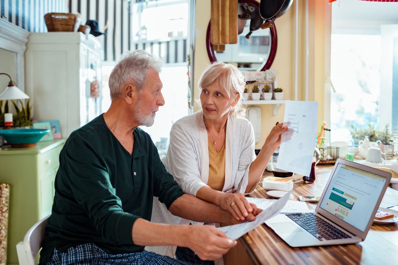 An older couple sat at a table looking at bills on paper and a laptop