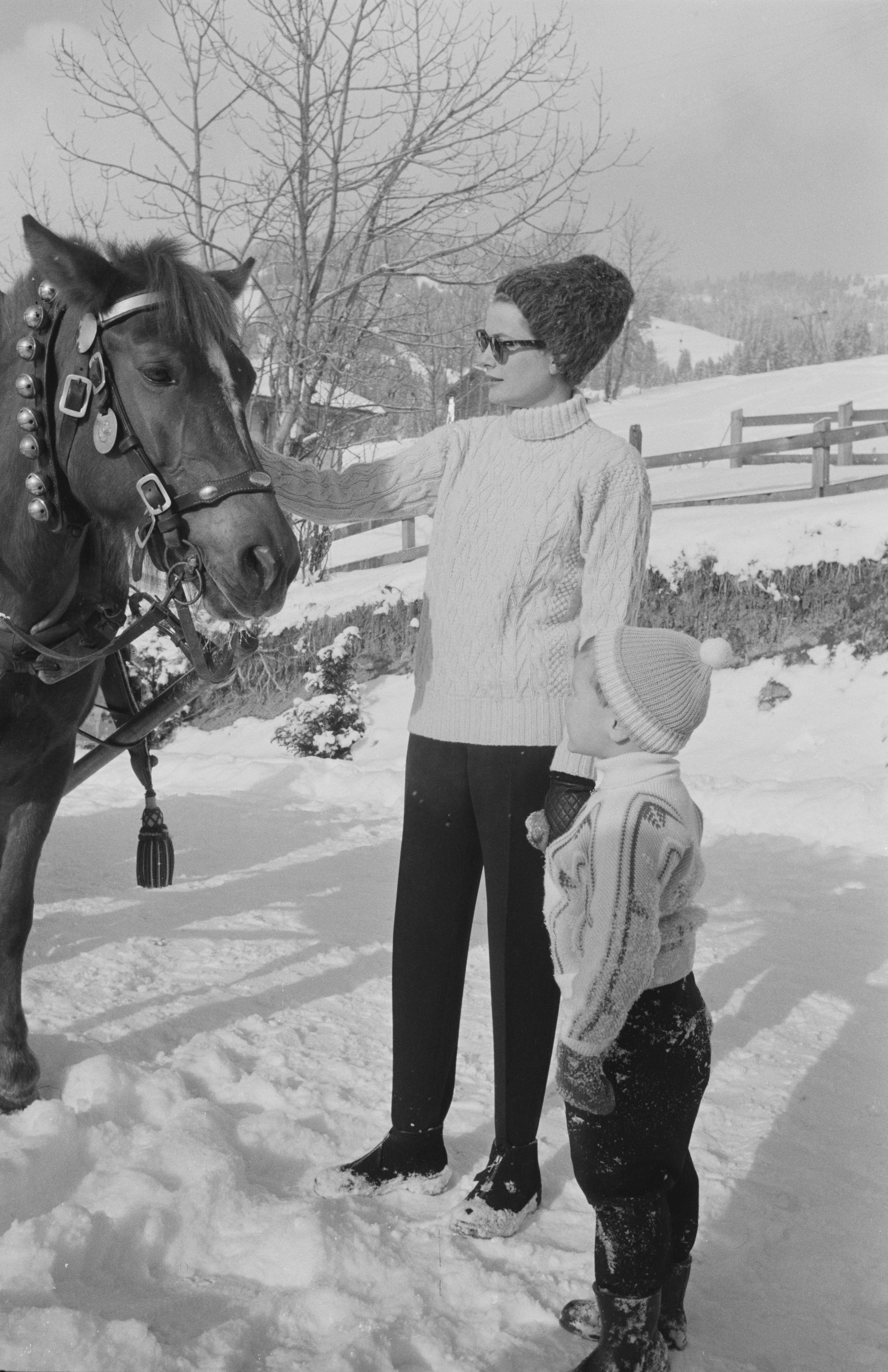 Grace Kelly and young Prince Albert wearing sweaters and winter hats in the snow petting a horse