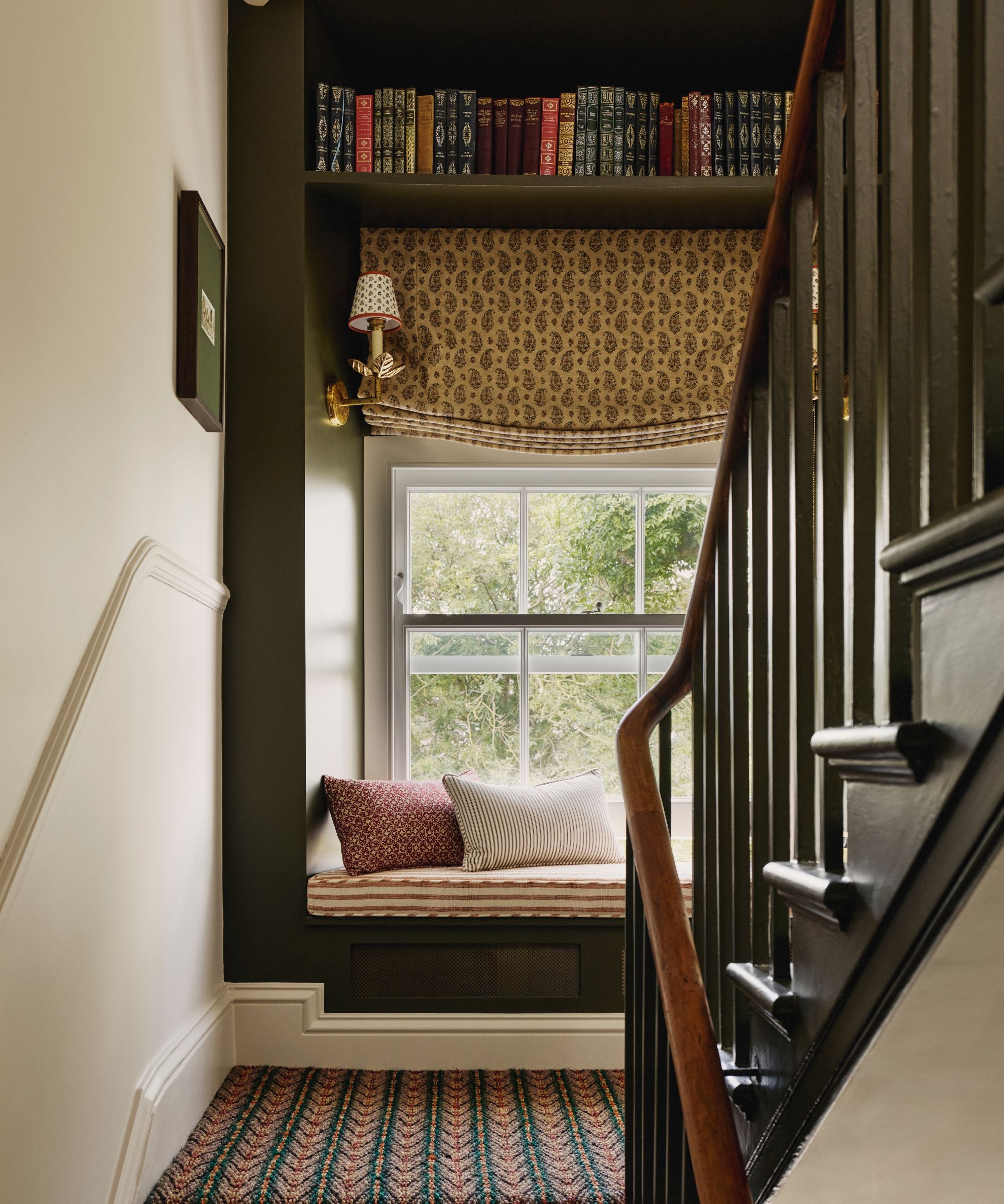 A landing with a dark green staircase and patterned carpet, a window seat featuring a striped cushion on a bench, a paisley Roman blind, a shelf of books above, and a wall sconce.