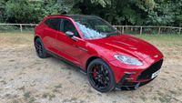 A red Aston Martin parked on a dirt path in front of a thin fence