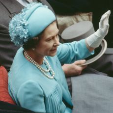 Queen Elizabeth wearing a blue hat and dress in a carriage waving