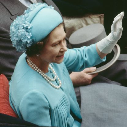 Queen Elizabeth wearing a blue hat and dress in a carriage waving