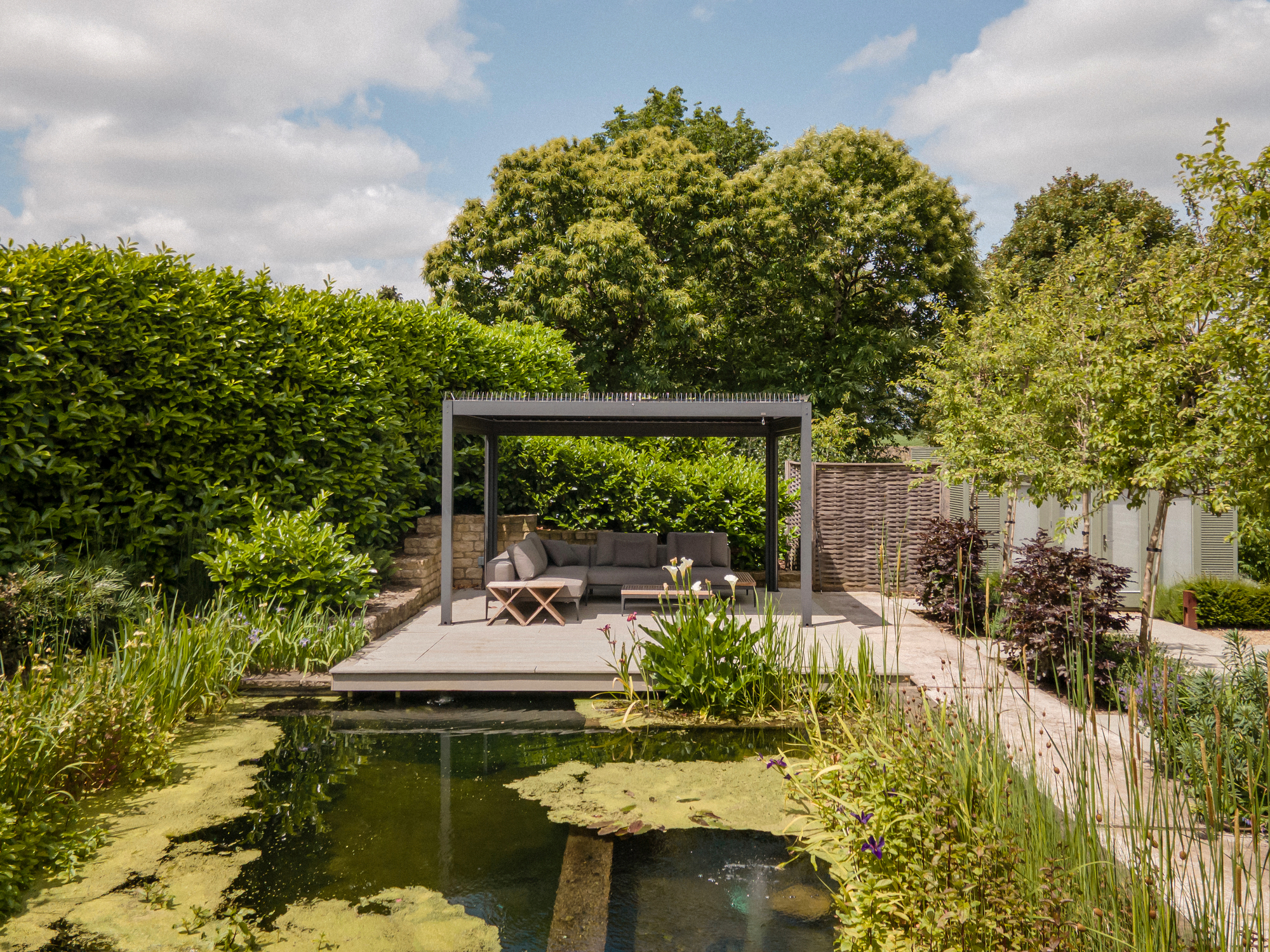 Waterside pergola on a deck by a large natural pond, surrounded by greenery