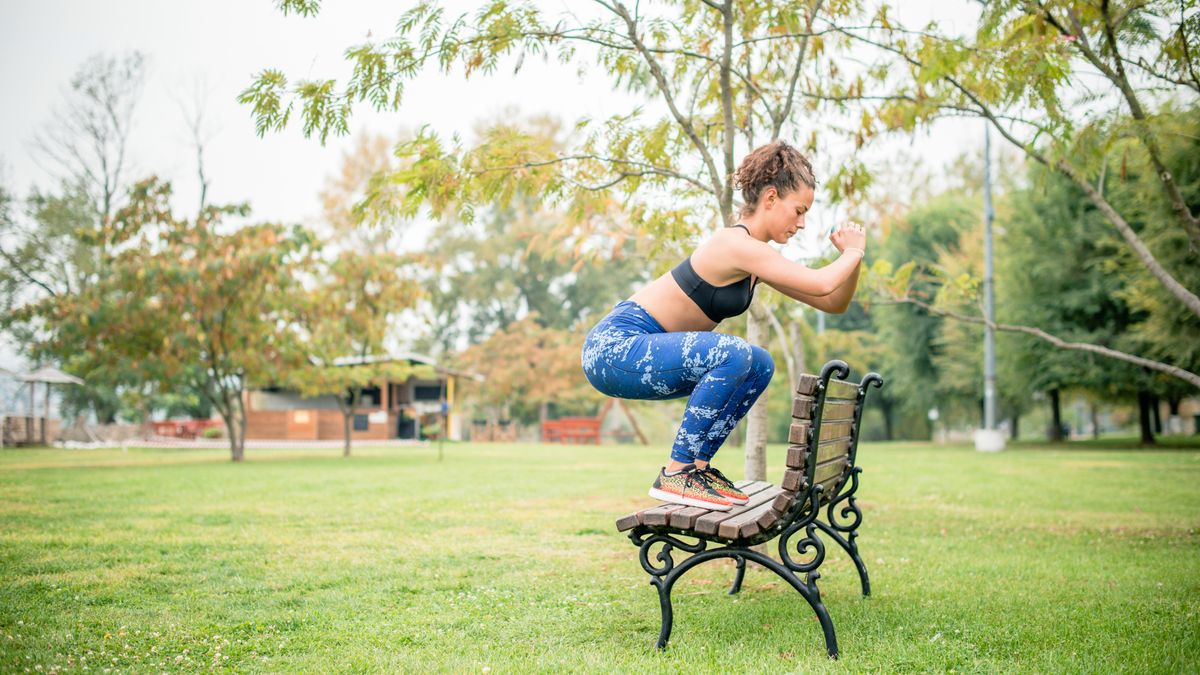 A woman doing a jump squat onto a bench in the park