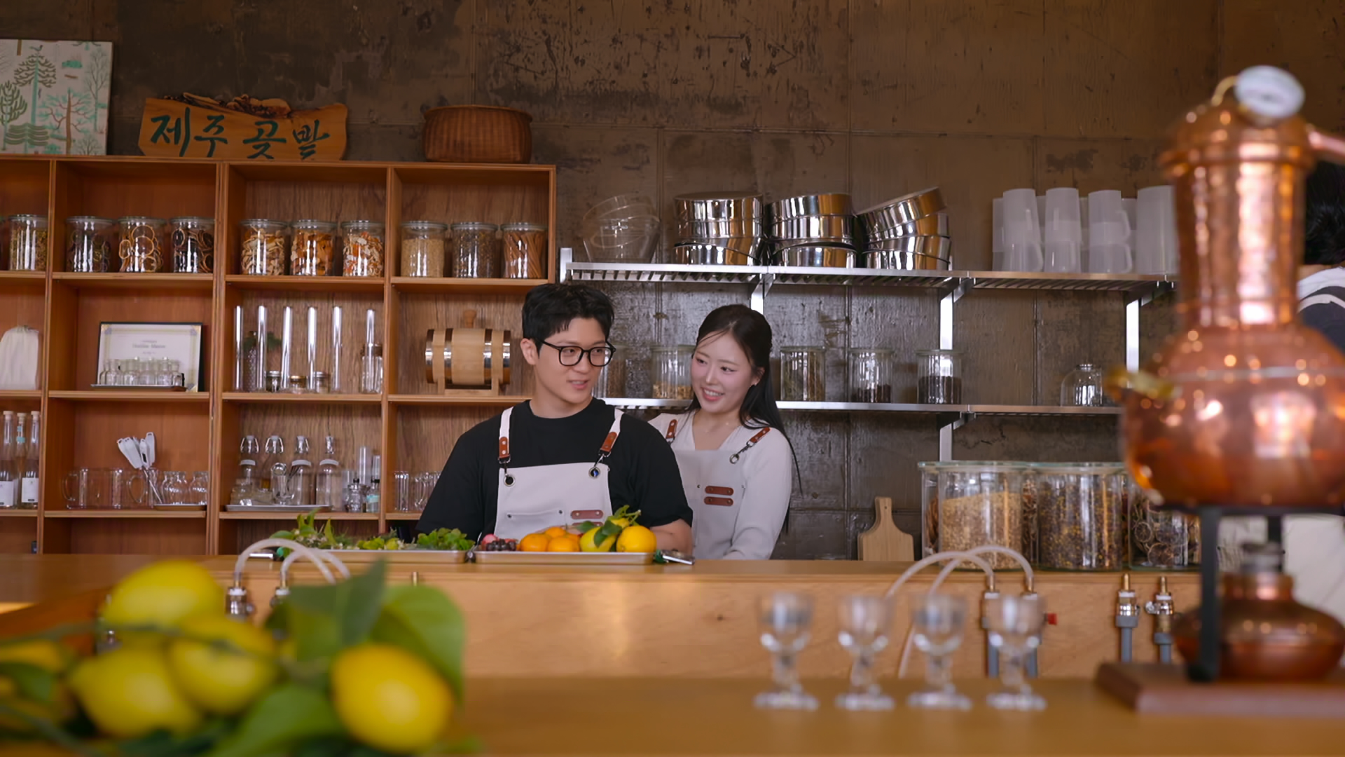 A Korean man and woman wear aprons while standing behind a bar in a restaurant kitchen, in Netflix's Korean reality show 'Better Late Than Single' season 2.