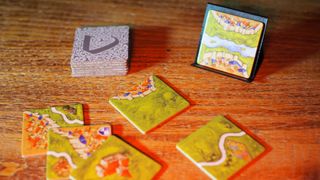 Carcassonne tiles laid out on a wooden table, with some stacked and one stood up against a black stand