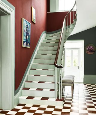 Red hallway with red a white checkered flooring and stairs, two pieces of artwork on wall beside stairs, rounded floral artwork on dark gray painted wall, upholstered bench beside stairs