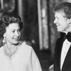 A black and white picture of Queen Elizabeth wearing diamond jewels and a white dress and Jimmy Carter wearing a tux at Buckingham Palace