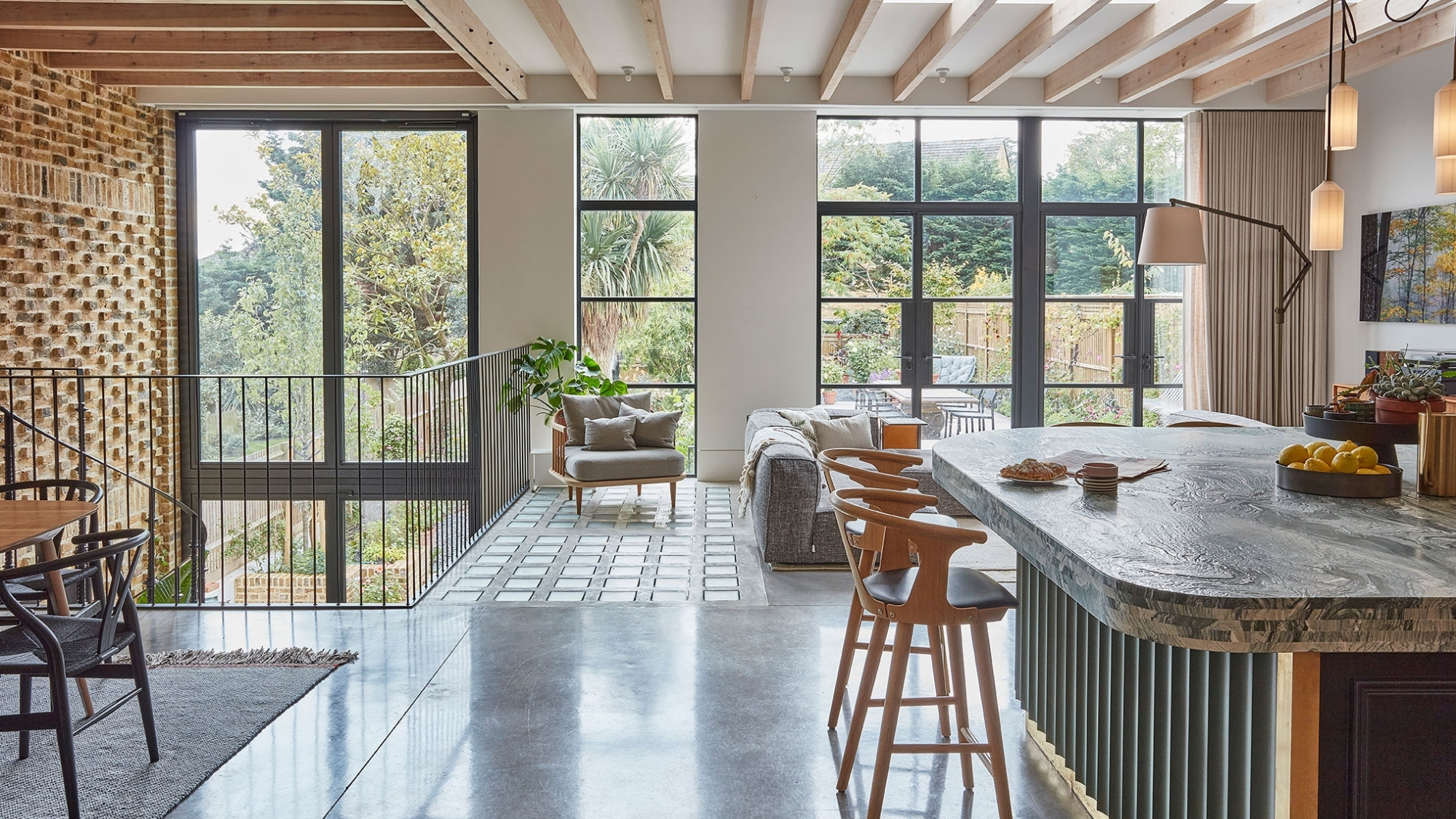 Marble kitchen island with fluted base, wooden bar stools, glazed doors overlooking a leafy garden view.