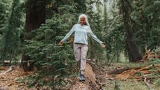 A woman walks along a fallen tree in a forest, smiling and holding her arms out to her sides. We see several large trees behind her.