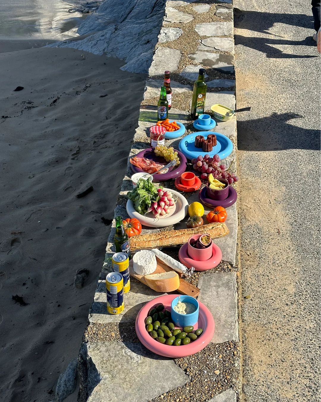 A series of colorful ceramic plates with a chunky design, sitting on a cement rim near a beach alongside cans of lemonade, baguette, and cheese, and filled with snacky bites.