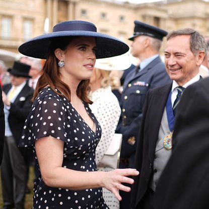 Princess Eugenie wearing a navy polka dot dress and blue hat talking to men at a garden party
