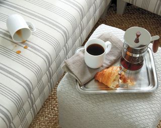 A cream striped upholstered sofa with mug and coffee stain next to a cafetiere and croissant pastry