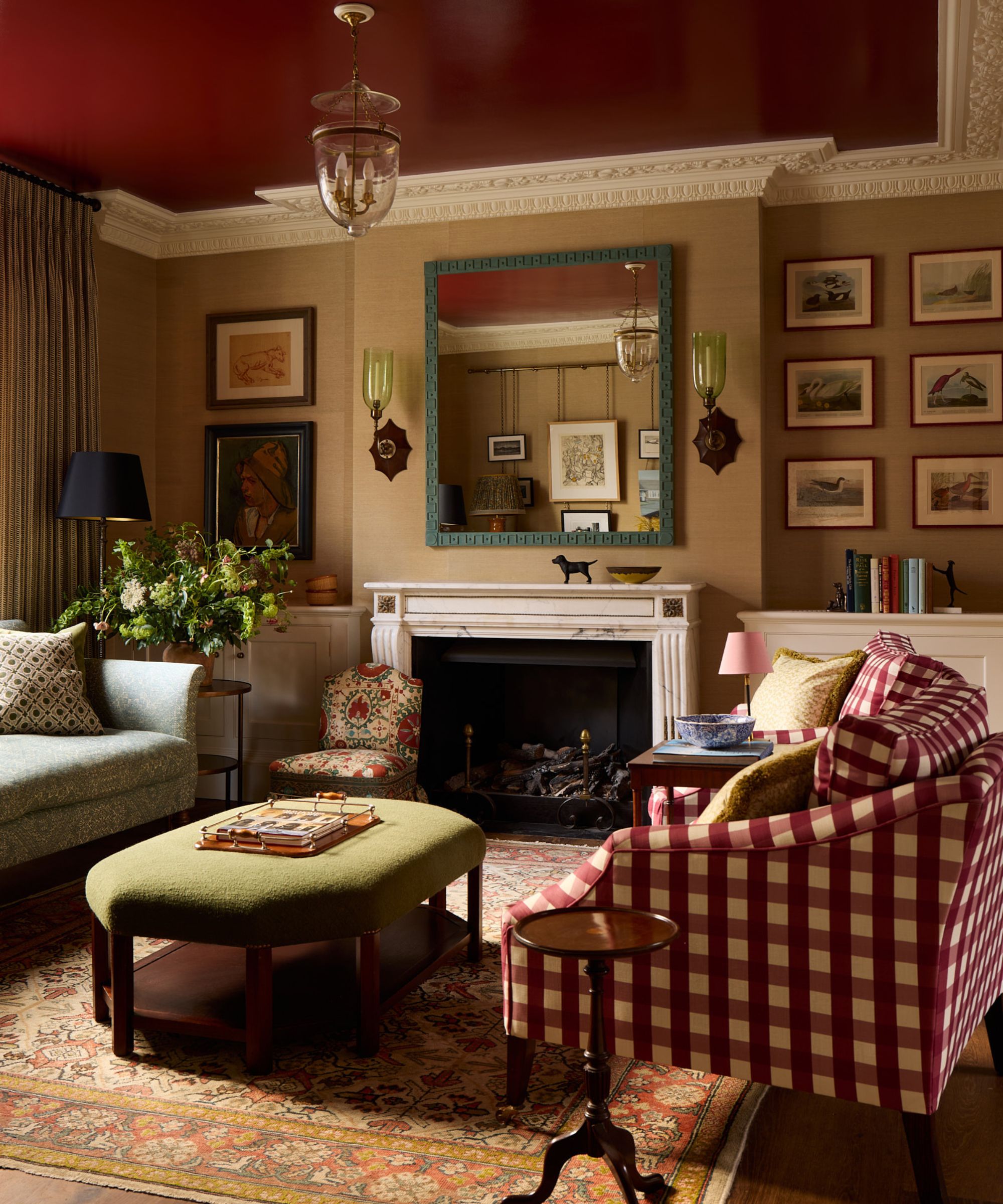 Colourful living room with red gloss ceiling, beige grasscloth walls, a marble fireplace, a blue mirror and two glass sconces either side, two red gingham armchairs, and a green velvet ottoman.