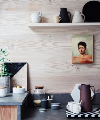 Corner of a kitchen with portrait artwork, high shelf with jugs, light wood wall, black stone countertops, kitchenware, ornaments in natural, neutral color palette on counter