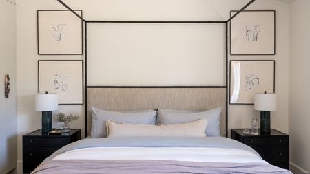 Bedroom with a black metal four-poster bed, lilac linen bedding and a neutral lumbar pillow, four framed prints on the wall in black frames, and black nightstands on either side with table lamps.
