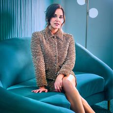 Melissa Fumero sits on a blue velvet couch wearing a textured collared jacket with matching skirt. 