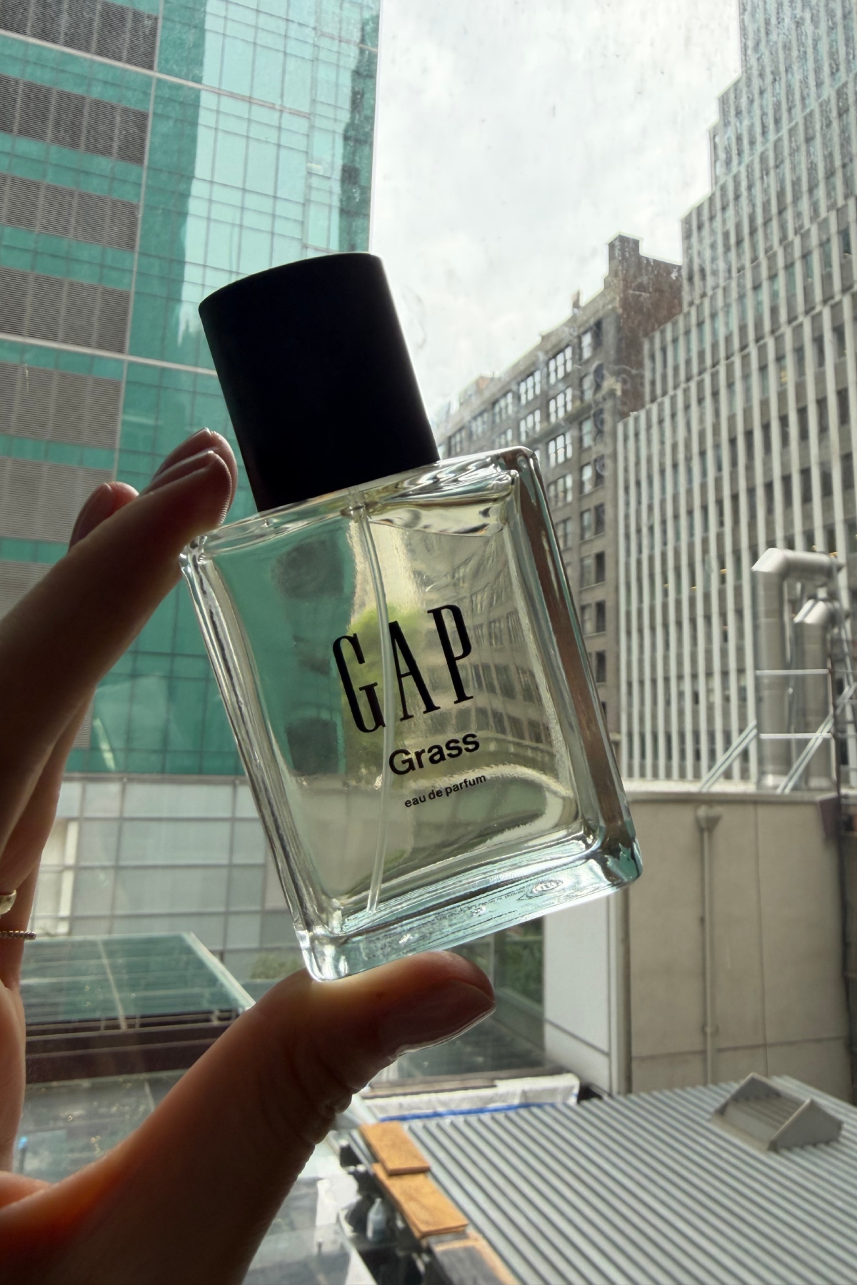 a hand holding up a clear glass bottle of Gap Grass fragrance to the window