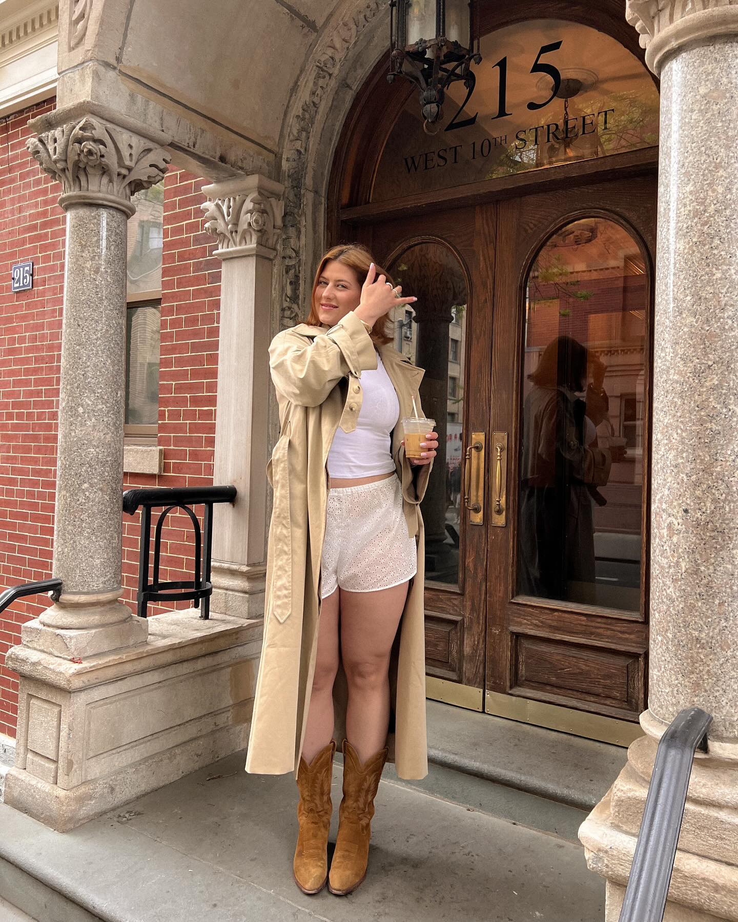 Raeaan wears bloomer shorts, cowboy boots, a white tank, and trench coat.