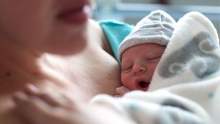 newborn wearing a tiny beanie leans against his mother's chest with his eyes closed and mouth yawning as both mother and baby are covered by a hospital blanket