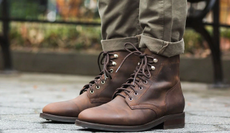 A picture of a person wearing brown leather boots from Thursday Boot Company and olive green trousers standing on a sidewalk in front of a bench.