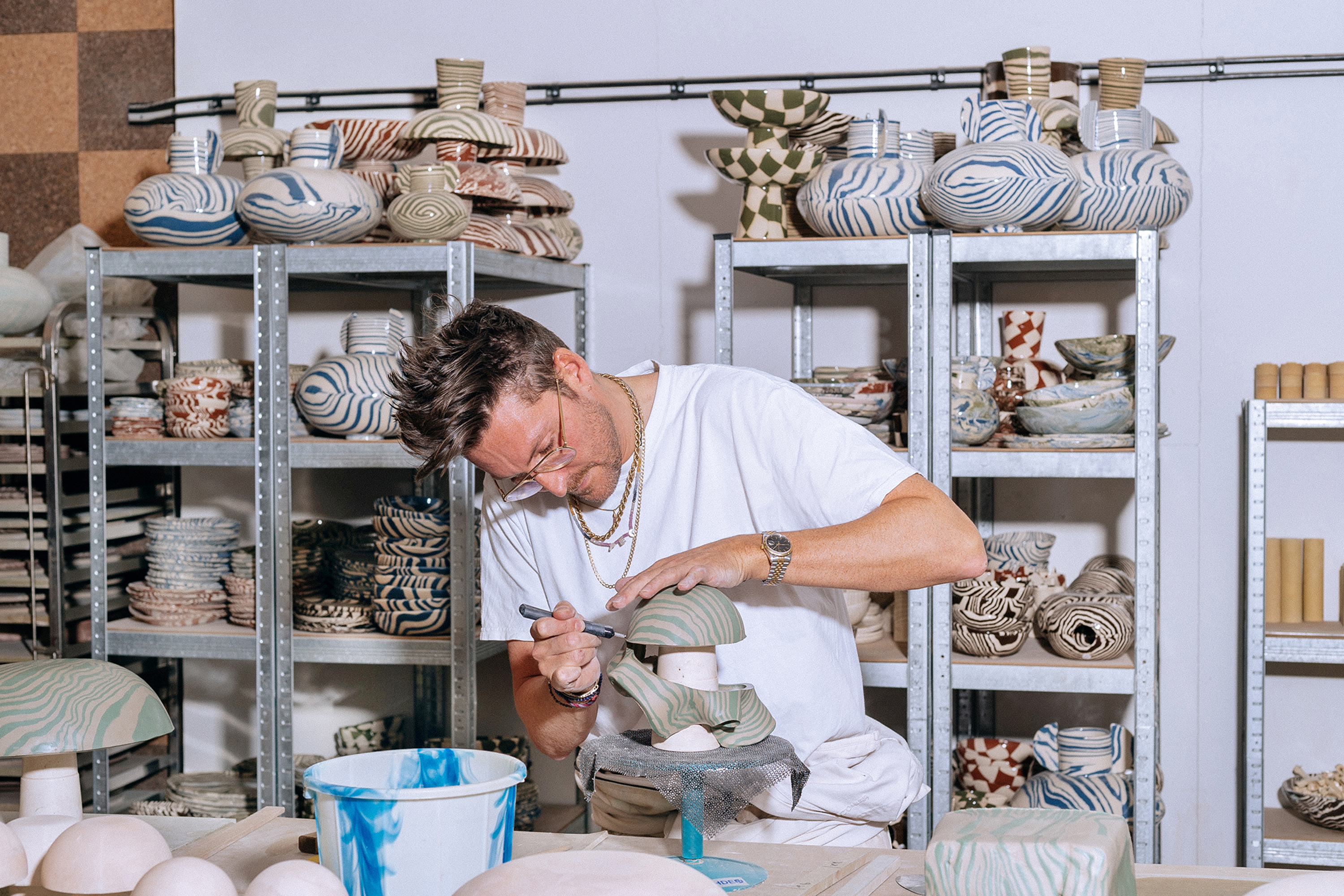 Henry Holland sculpting a piece of clay in his studio with shelves of ceramics behind him