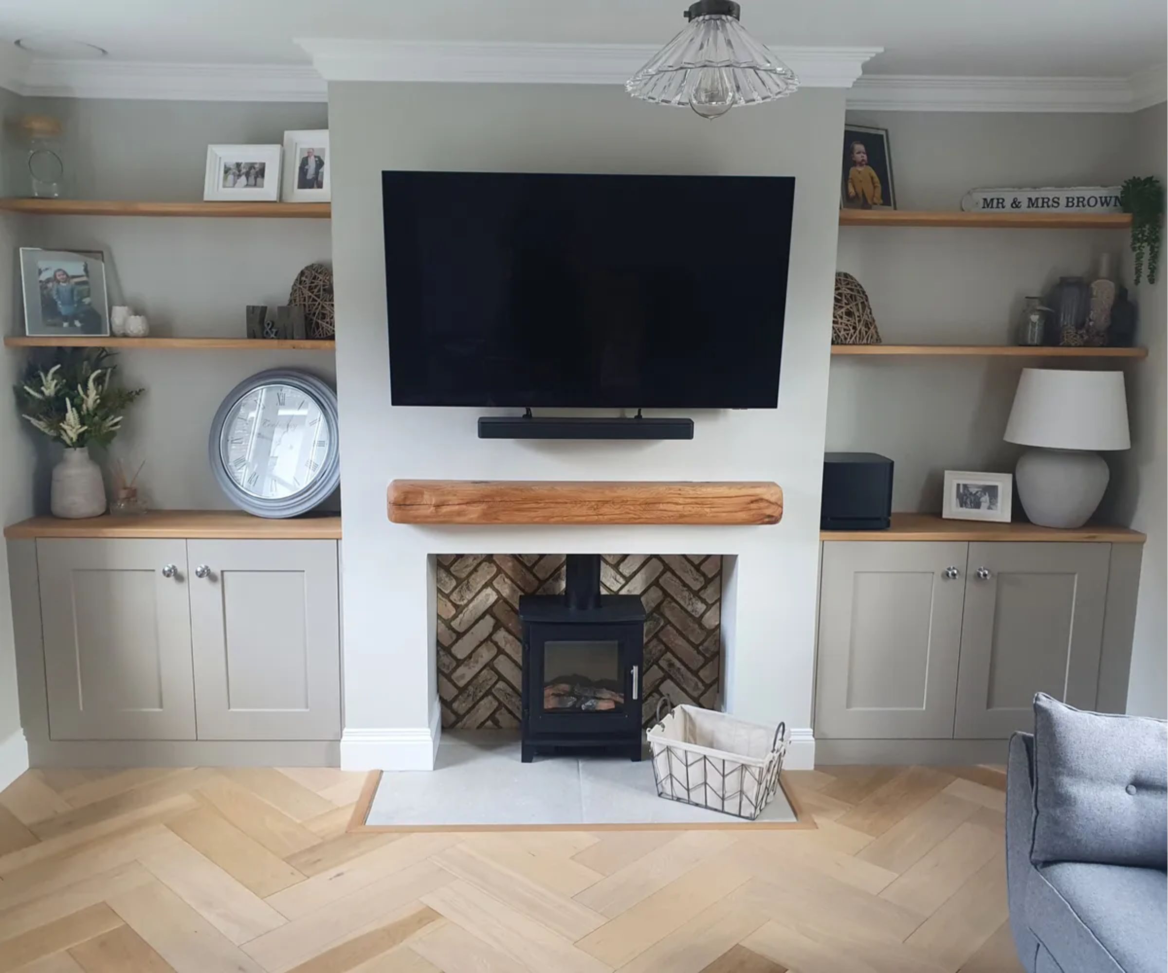 A small log burner with a rustic oak mantel is set into a herringbone brick chimney breast, with shelves and cabinets on either side.