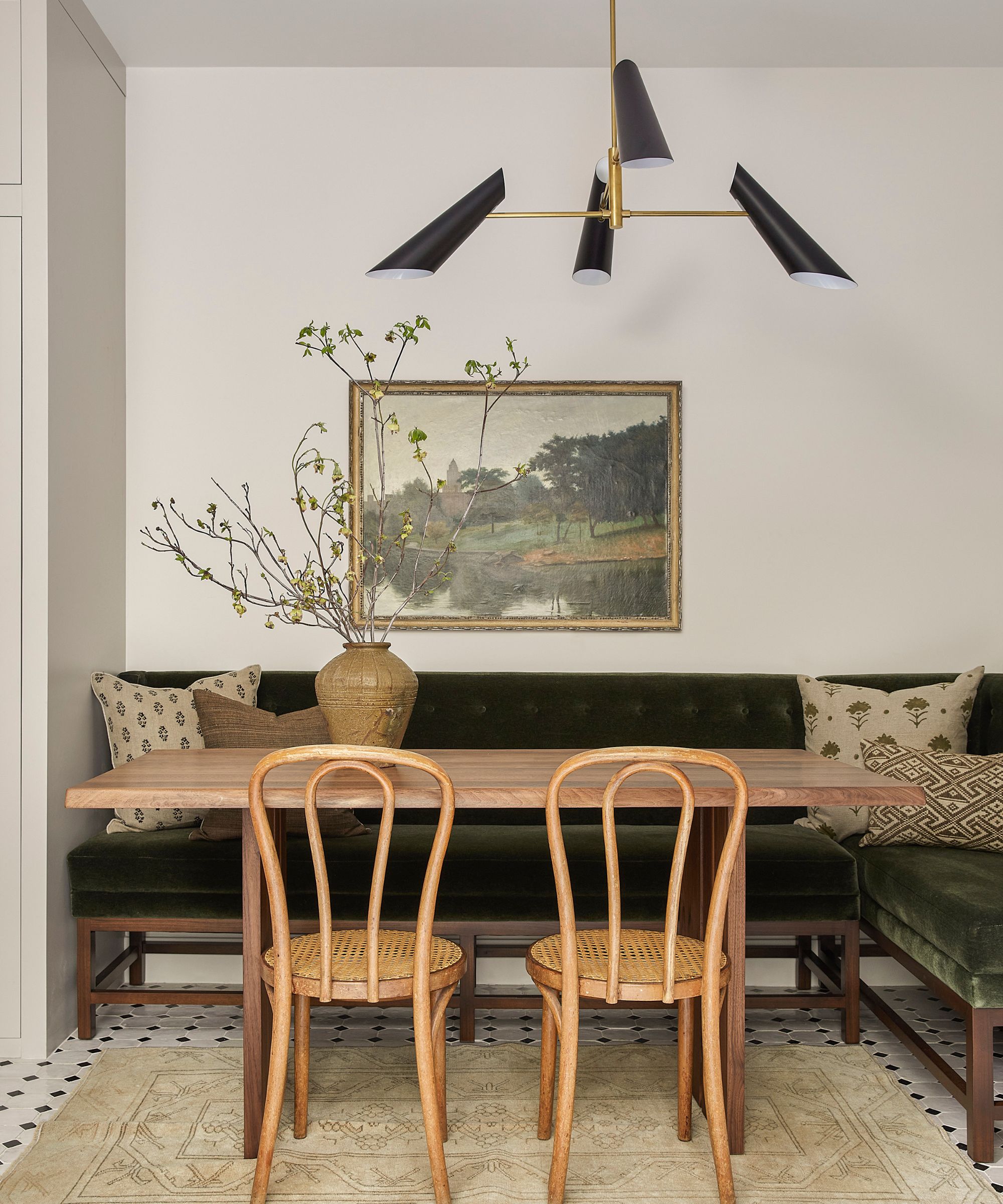 Dining nook with a long wooden table, deep green velvet banquette, rattan chairs, and a modern chandelier.