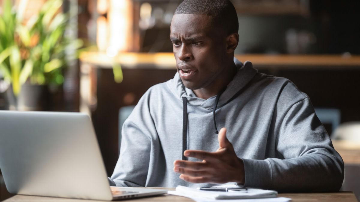 A man at a desk using a laptop and holding his hands up, while having a confused look on his face